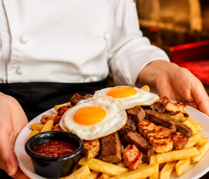 Plato de papas fritas con carne, pollo a la plancha, cebolla caramelizada, huevos fritos y salsa, presentado por un chef de L'angolo.